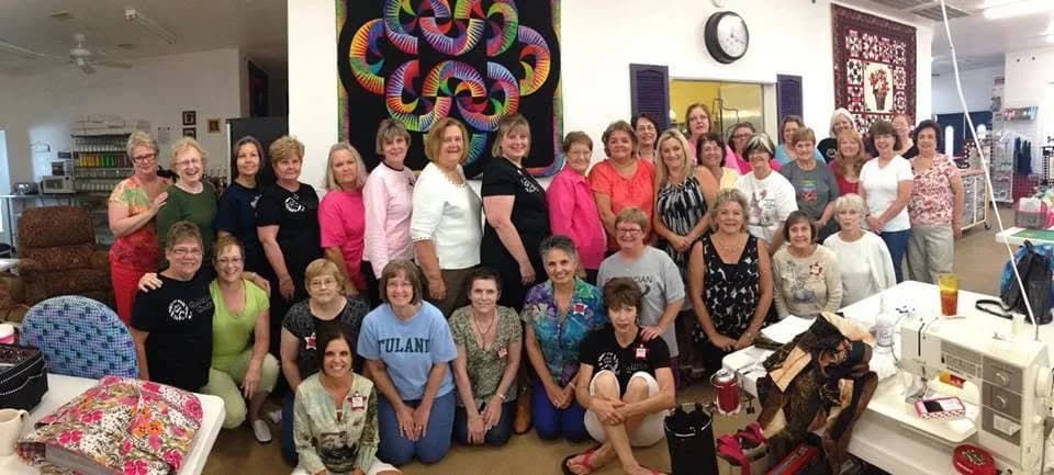 Large retreat group posing in the workroom with quilts behind them